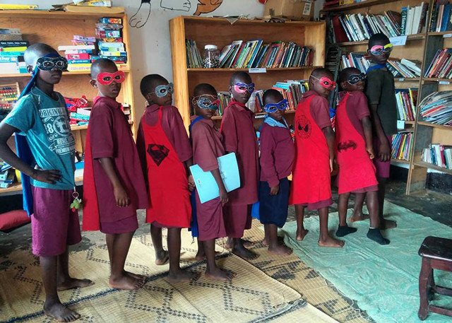 Children stand in a line wearing super-hero capes and masks made from cereal boxes, making bold super-hero poses.